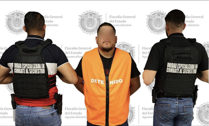 Image: A man in an orange vest faces the camera as two men with their backs turn are beside him.