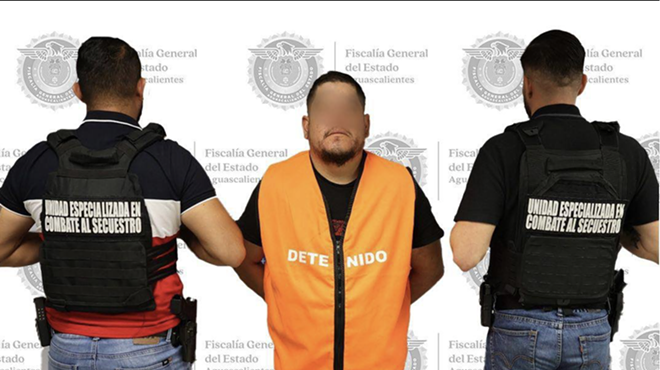A man in an orange vest faces the camera as two men with their backs turn are beside him.
