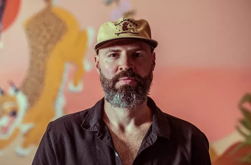 Image: man with beard in a flat brim hat and button down shirt in front of a pink background