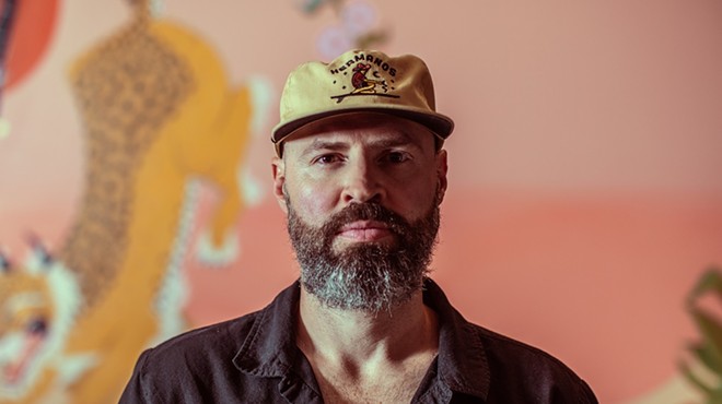 man with beard in a flat brim hat and button down shirt in front of a pink background
