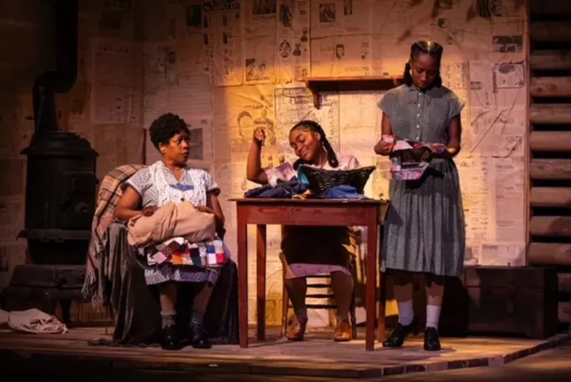 Image: Two women sit, and one stands around a wooden table.