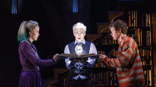 A man with silver hair holds a book, which two people grasp on either side.