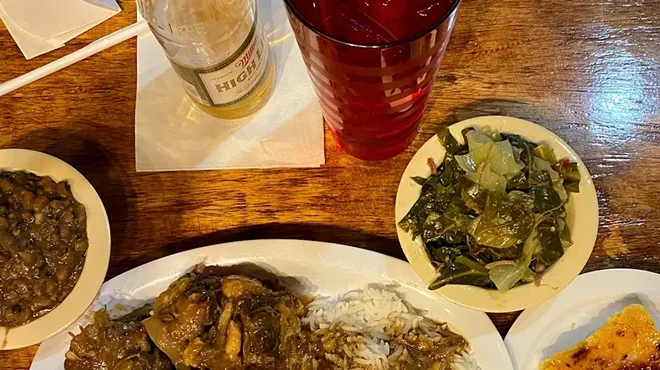 a beer on a table with a plate of oxtails and rice and a bowl of collard greens on the side.