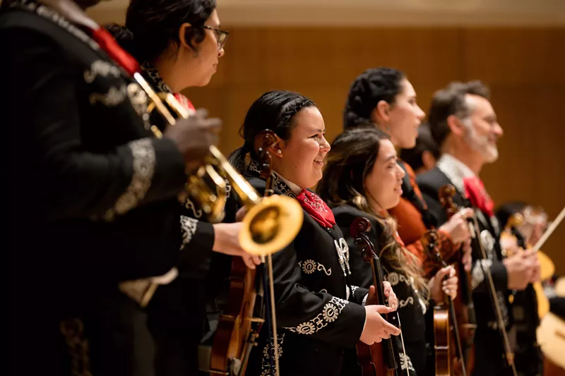 Image: 6 mariachi musicians stand on a stage while facing a crowd of supporters and fans.