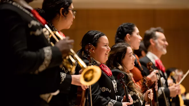 6 mariachi musicians stand on a stage while facing a crowd of supporters and fans.
