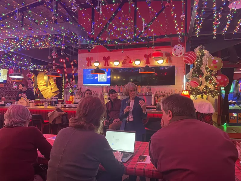 Image: A woman in a blue suit speaks to people gathered in a room decorated for Christmas.