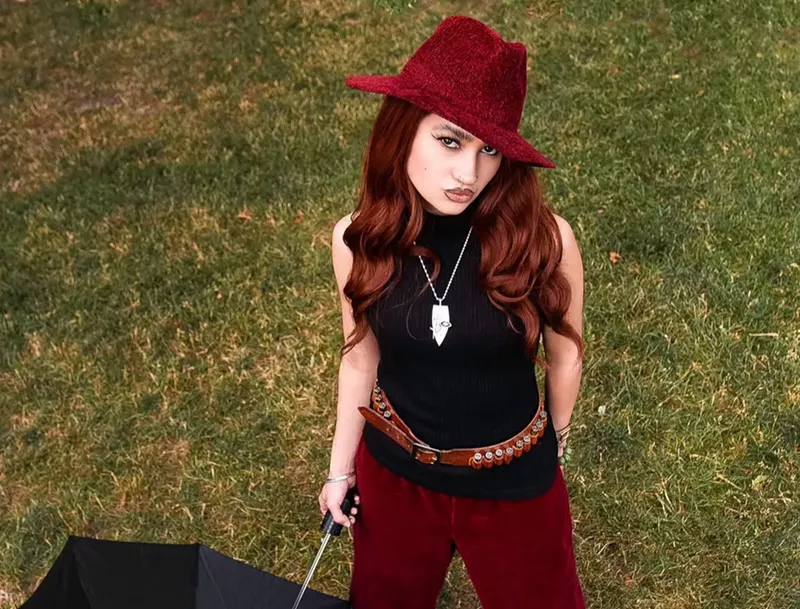 Image: Kitty wears a red suede hat and pats with a black top while holding an umbrella on a lawn.