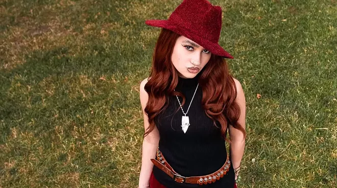 Kitty wears a red suede hat and pats with a black top while holding an umbrella on a lawn.
