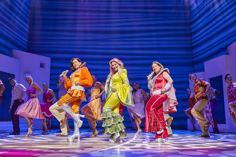 Image: women in orange, yellow and red costumes dance on stage with blue background