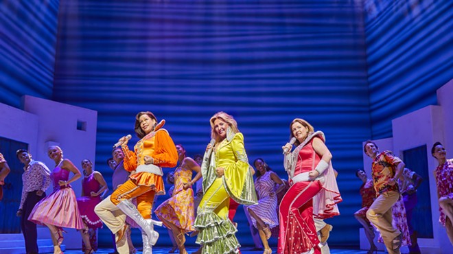 women in orange, yellow and red costumes dance on stage with blue background