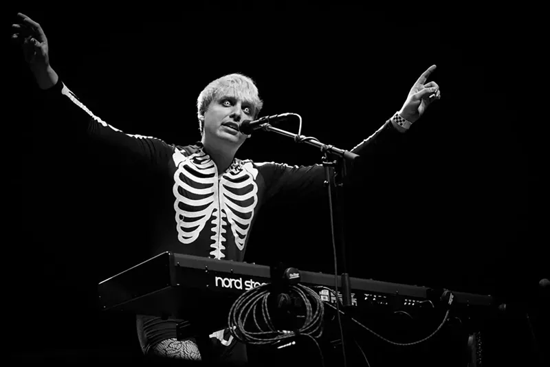 Image: man in skeleton sweater stands at a keyboard and mic with arms in the air