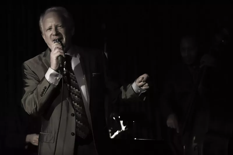 Image: A man in a suit sings with a microphone on stage.