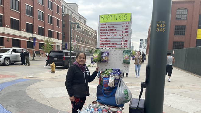 A woman stands on the corner with a cart containing ballpark snacks.
