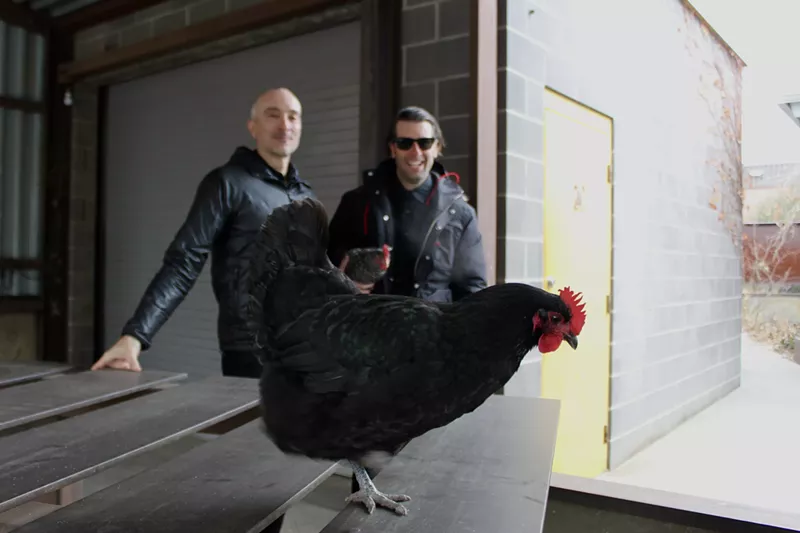Image: Two men stand behind a black chicken on a table