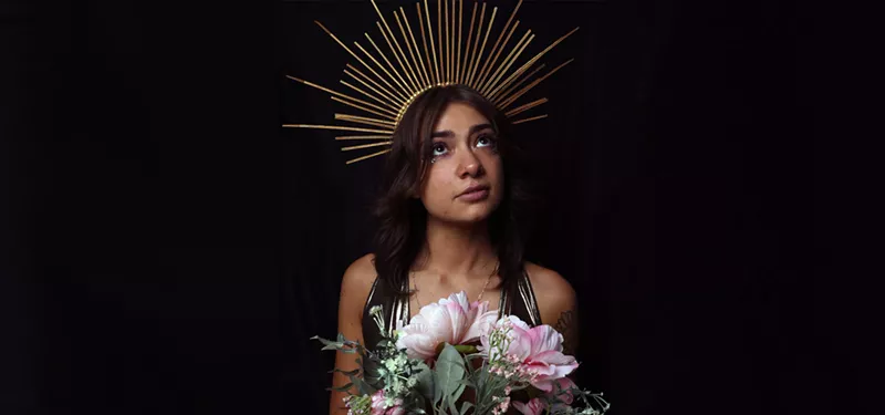 Image: brunette woman wearing gold crown and holding pink flowers.