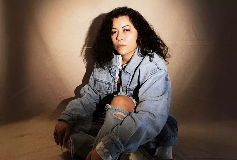 Image: A woman wearing a all-denim look and turquoise nose-ring poses in front of a beige backdrop for the camera.
