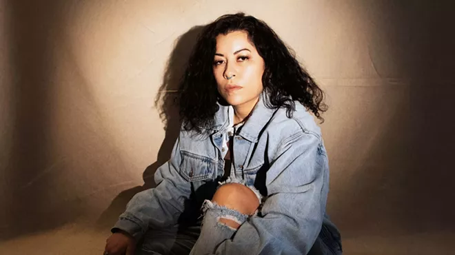 A woman wearing a all-denim look and turquoise nose-ring poses in front of a beige backdrop for the camera.