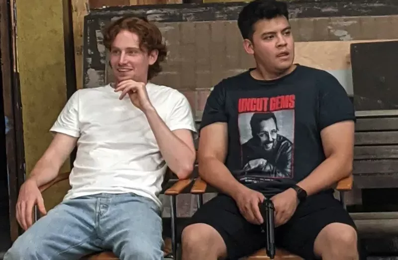 Image: a ginger boy in a white t shirt and a man with black hair in a black shirt holding a fake gun.