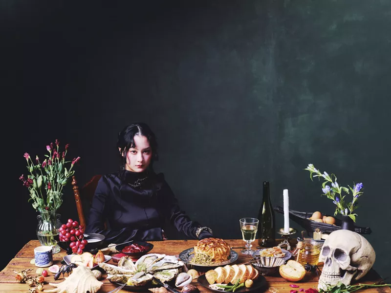 Image: Someone sitting at a dinner table with a skull and several food items on a dark green-blue background