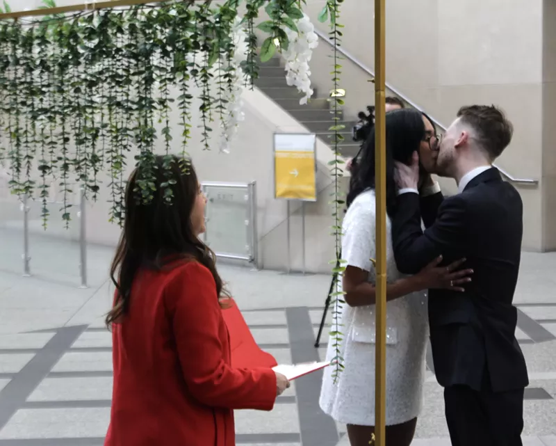 Image: A man and woman kiss at the altar, an officiant wearing red looks on
