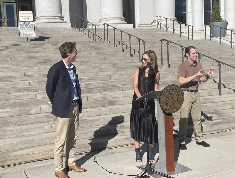Image: A man in a blue blazer and khakis converses with a woman wearing a long black dress behind a podium.