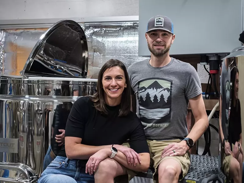 Image: a woman and a man sitting on step ladders in front of brewing equipment
