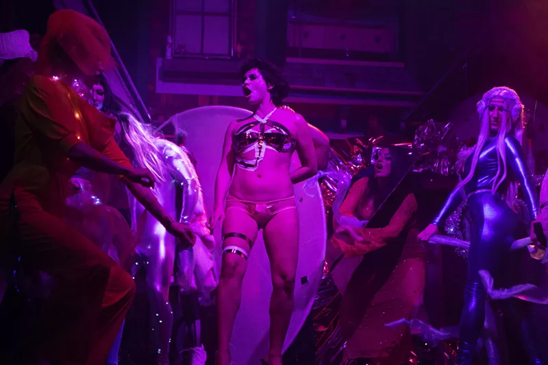 Image: drag performers on stage with purple lighting.