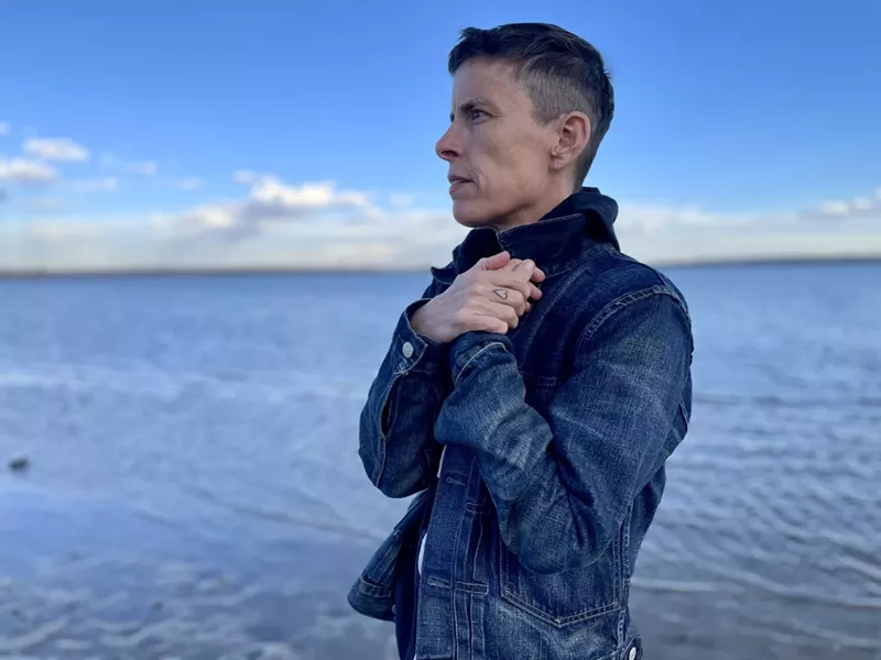 Image: person with short hair has hands to the heart as they stand in front of water