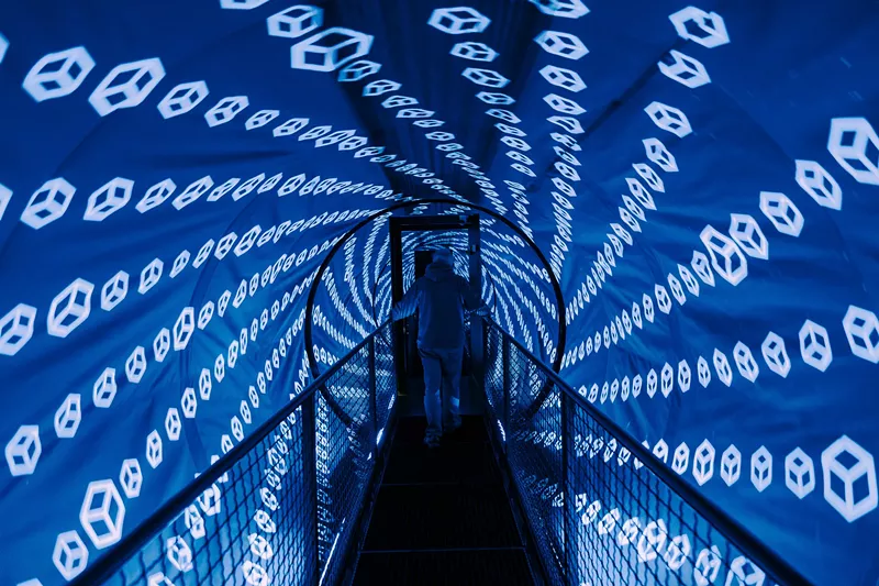 Image: blue lights cover a tunnel-shaped room with a bridge in the center