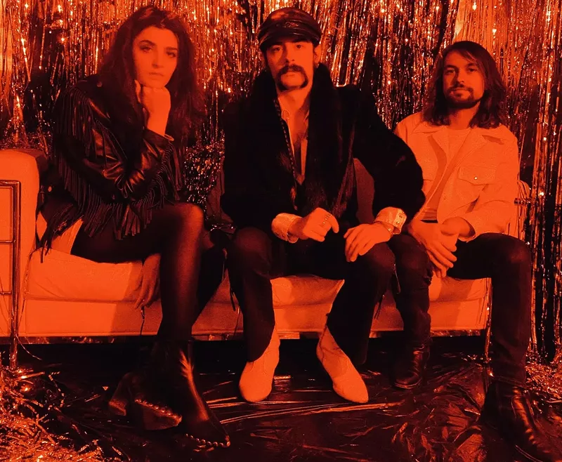 Image: three people sitting on a couch in front of a tinsel wall in orange lighting
