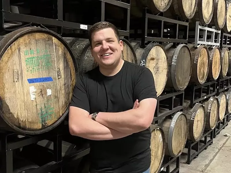 Image: a man in a black t shirt stands in front of wooden barrels