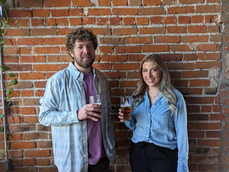 Image: A man and a woman stand in front of a brick wall.