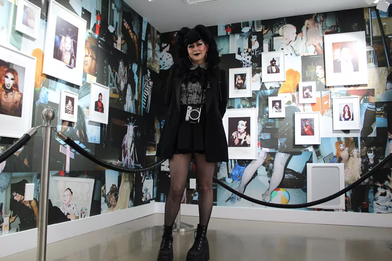 Image: A woman dressed in black stands in front of a wall of photos