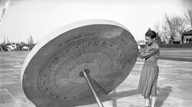 An old photo of a woman standing by a sundial