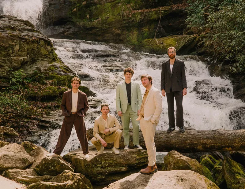 Image: Four men standing, one squatting, on top of rocks and logs above a running creek. All dressed in different color suits.