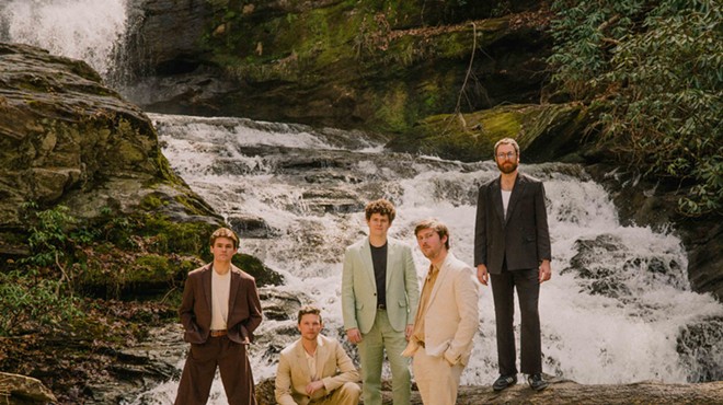 Four men standing, one squatting, on top of rocks and logs above a running creek. All dressed in different color suits.