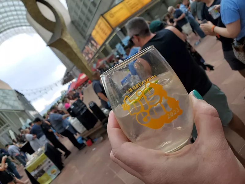 Image: hand holding a glass in front of people standing near a statue