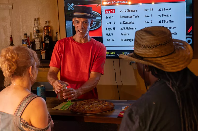 Image: man in red shirt, black hat, in front of menu.