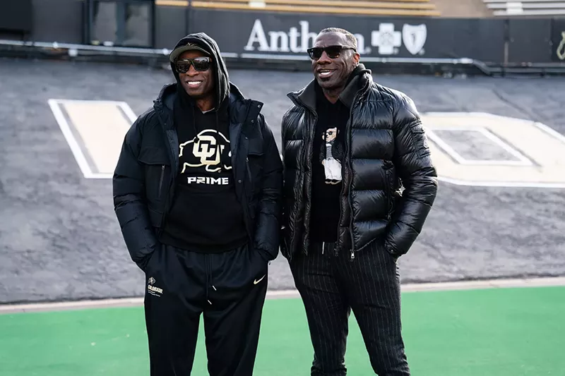 Image: Two Black men in black coats and pants stand together smiling.