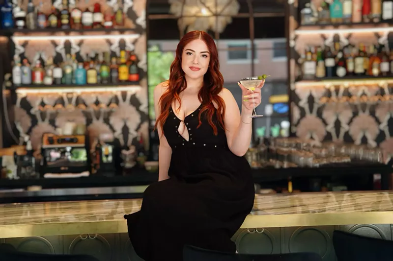 Image: woman n a black dress sitting on a bar and holding a martini