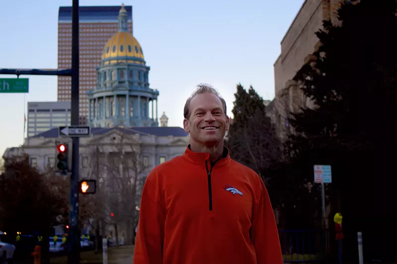 Image: Jason Clark wearing an orange Broncos sweatshirt standing in front of the Colorado Capitol building.