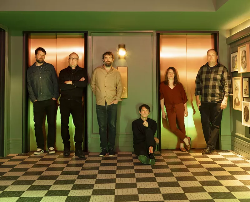 Image: The members of the band Cursive in a hotel's elevator bay, all standing except songwriter, guitarist and vocalist Tim Kasher.