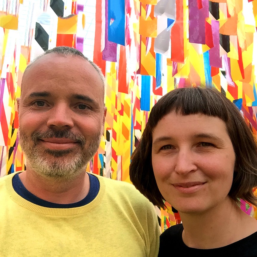 man in yellow shirt and woman with bangs in front of colorful art installation