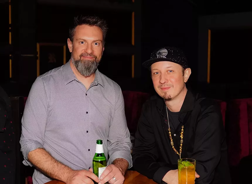 a man in a grey shirt holding a beer next to a man in a black hat