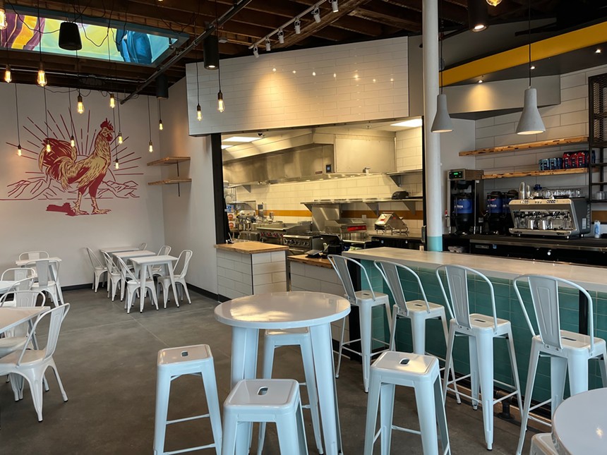 Restaurant interior with kitchen and bar and tables and white and teal decor with a chicken mural