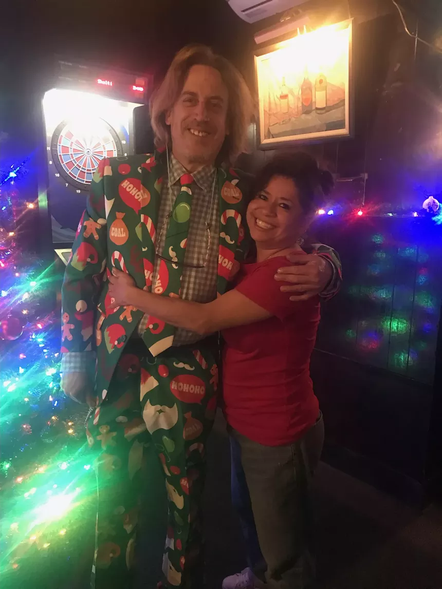 a man in a Christmas-themed suit hugs a women standing front of a Christmas tree
