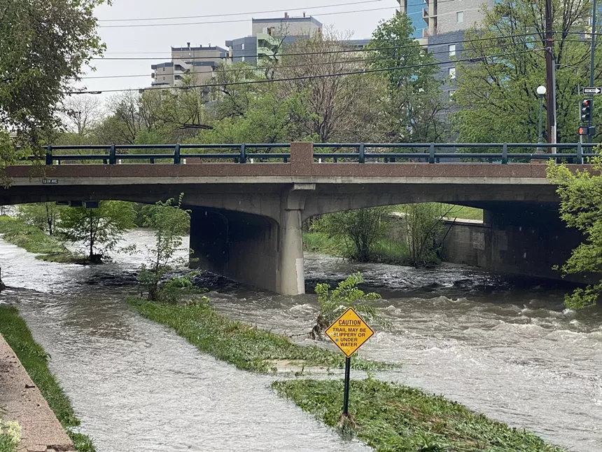 A bridge runs over a river which has overflowed onto an adjacent walking trail.