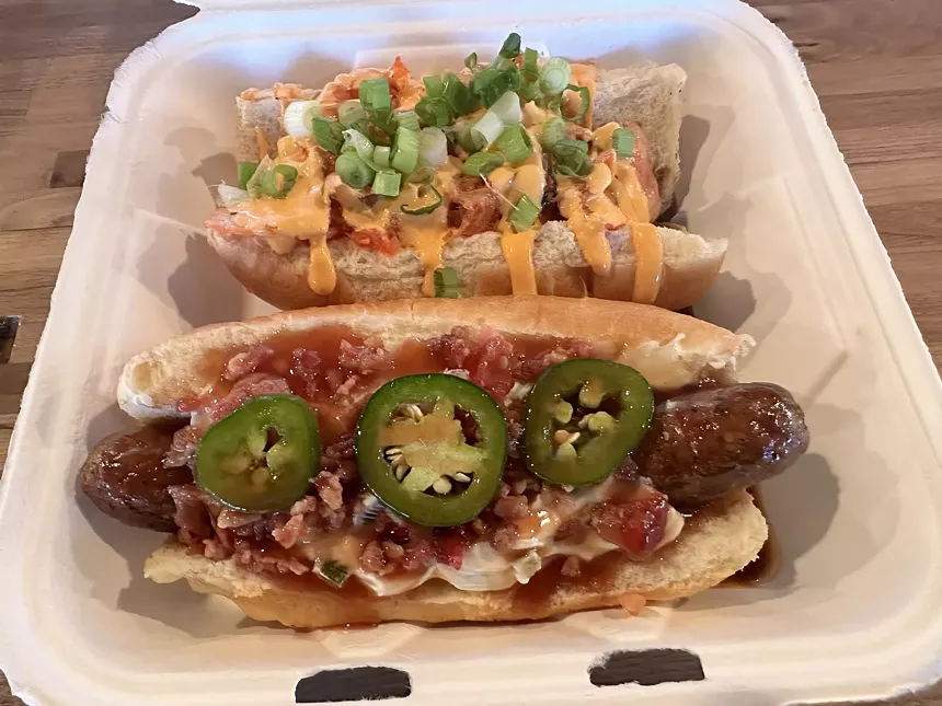 two hot dogs with various toppings in a to-go box
