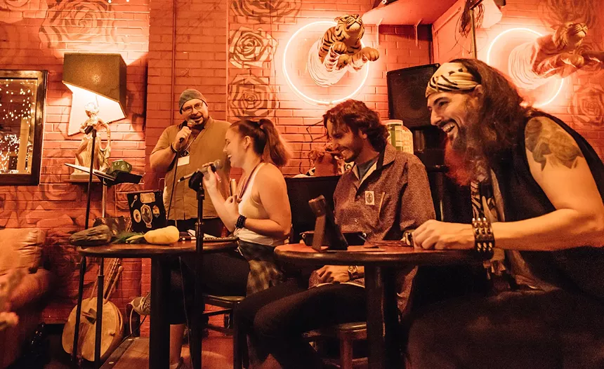 man speaks into a microphone to three people sitting at tables laughing