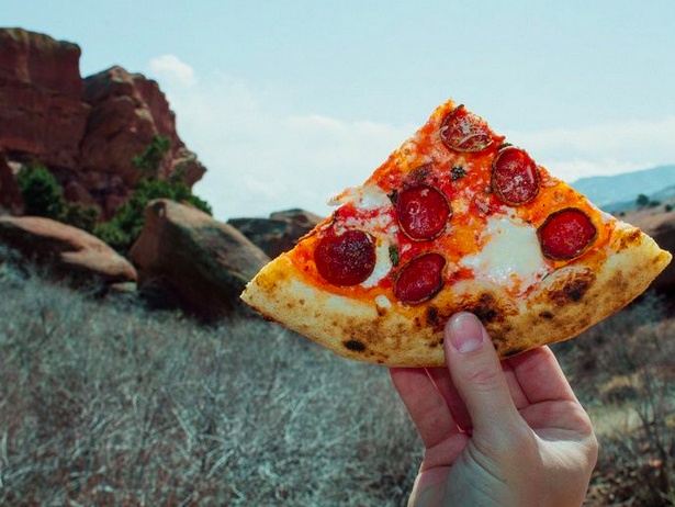 a hand holding a slide of pepperoni pizza with a view of rocks in the background